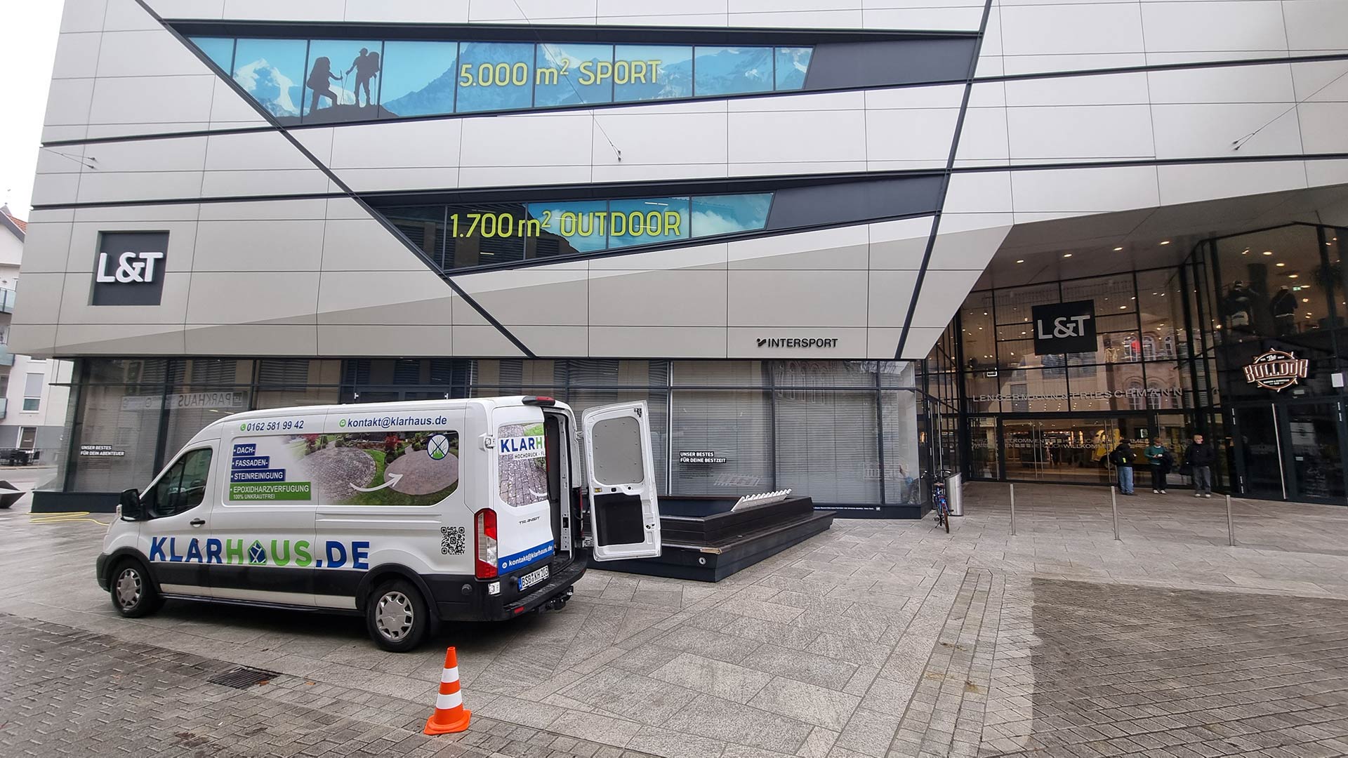 Ein weißer Lieferwagen steht vor einem modernen Gebäude mit großen Fenstern und Werbebeschriftungen.