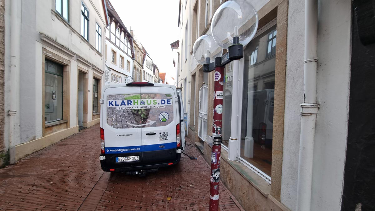 Ein weißer Transporter in einer engen Straße, umgeben von alten Gebäuden und einer Straßenlaterne.