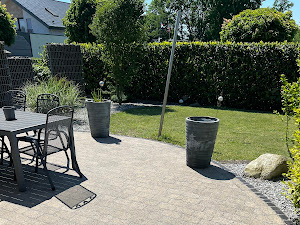 Ein Garten mit Tisch, Stühlen, Pflanzen in grauen Töpfen, grünem Rasen und Hecke im Hintergrund.