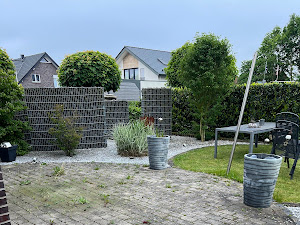 Ein gepflegter Garten mit modernen Gabionen, Kieswegen, Bäumen und einem Tisch im Freien.