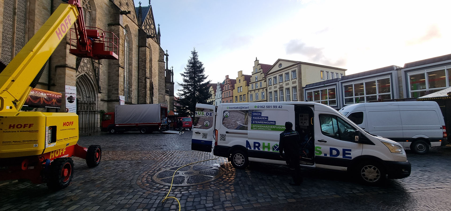 Ein Mann in dunkler Kleidung steht lächelnd neben einem weißen Transporter auf einem Marktplatz im Abendlicht