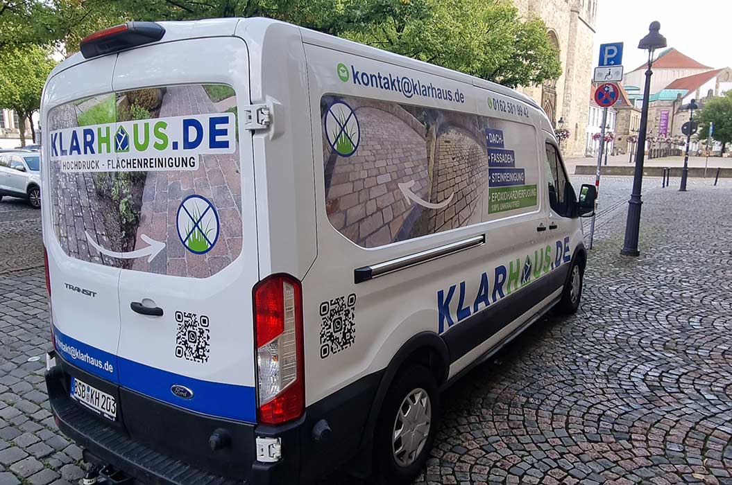 Ein weißer Firmenwagen mit blauer und grüner Werbefolie steht auf Kopfsteinpflaster