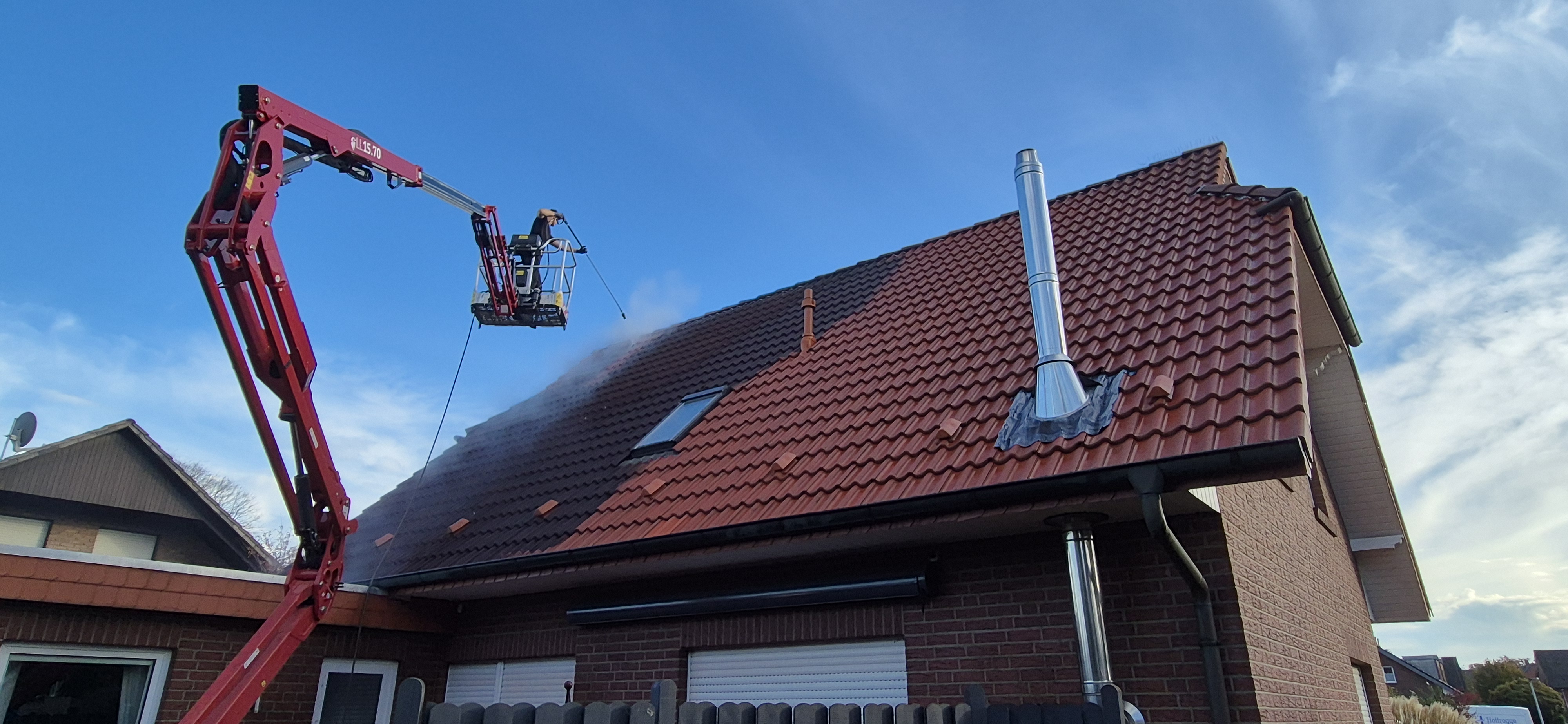 Eine Person mit Helm reinigt lächelnd das rot gedeckte Hausdach mit Wasserdampf