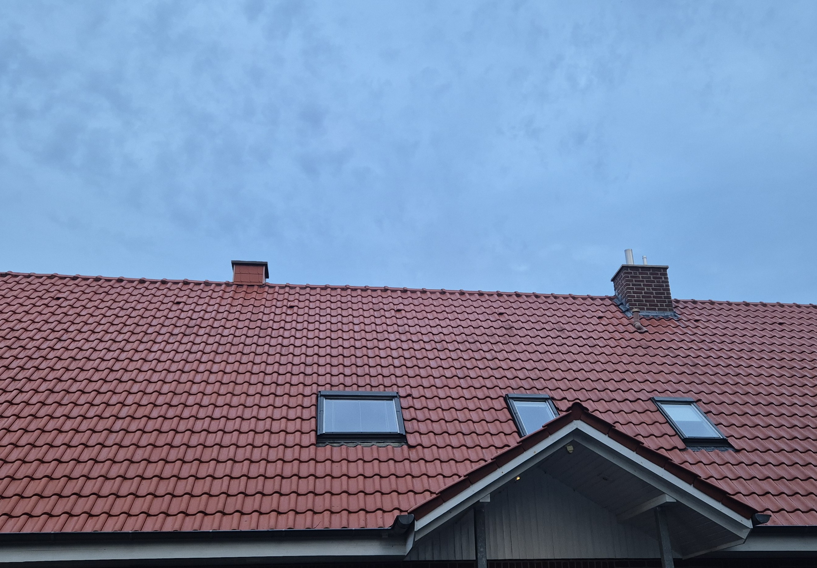Ein rotes Ziegeldach mit zwei Dachfenstern und einem gemauerten Schornstein vor blauem Himmel