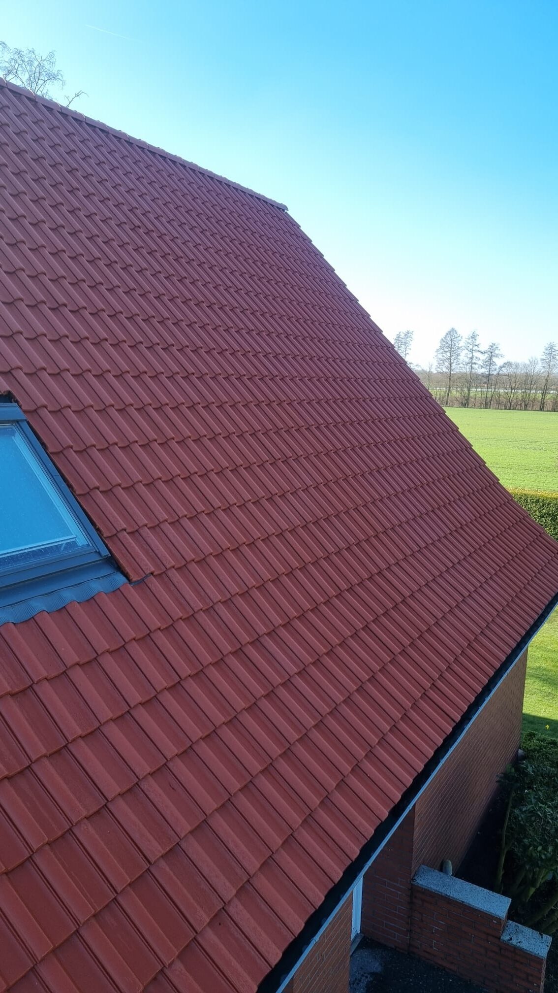 Ein rotes Ziegeldach mit Dachfenster vor blauem Himmel und grüner Landschaft