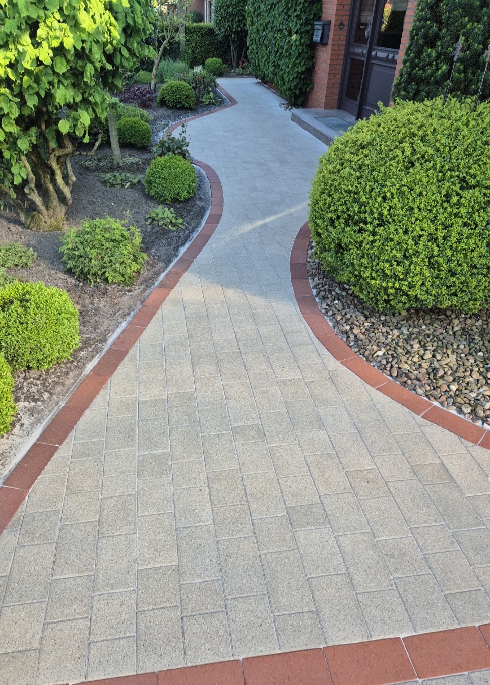 Ein geschwungener heller Weg mit roten Kanten führt durch einen grünen Garten