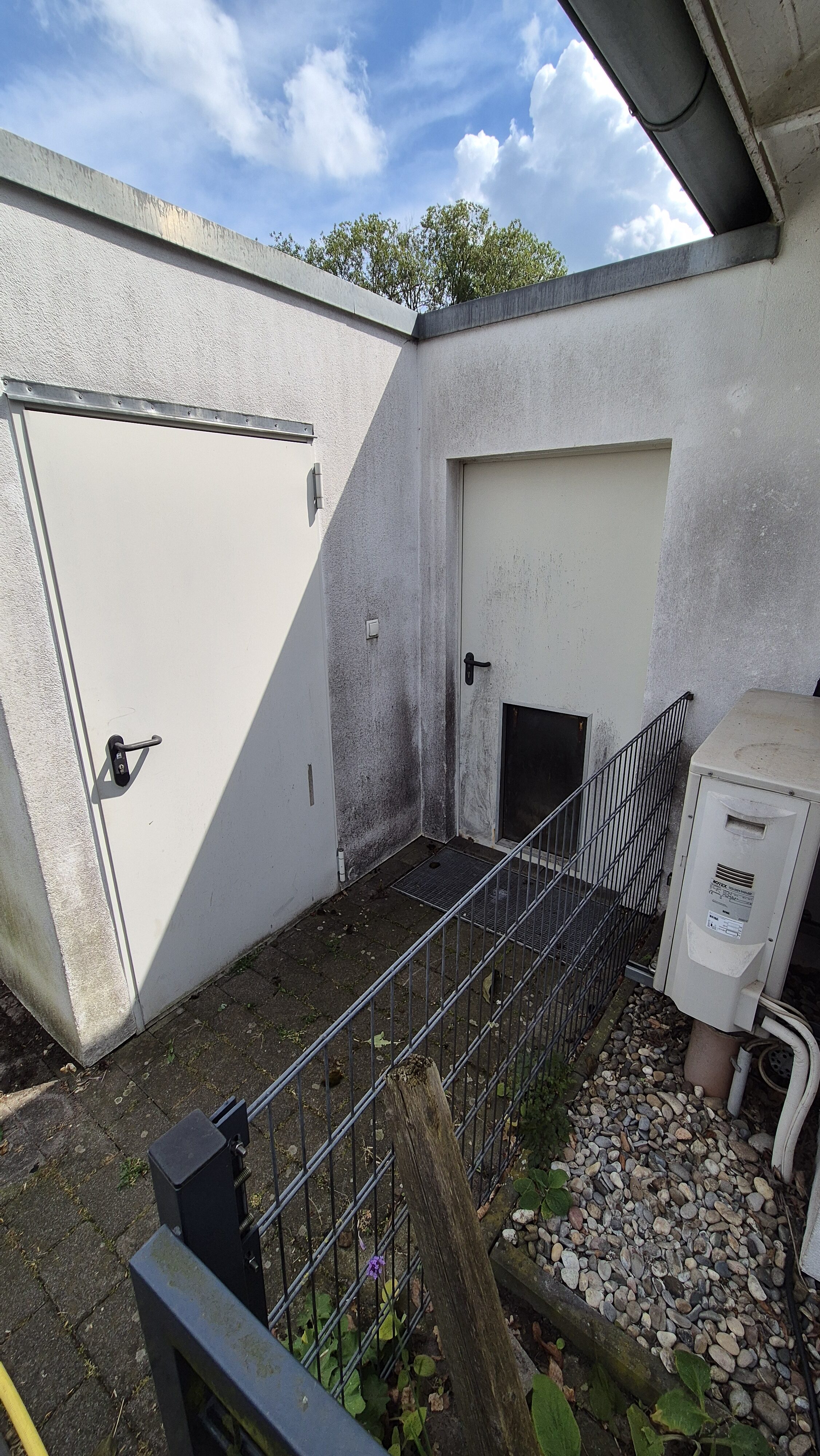 Ein kleiner Hof mit weißen Türen, einer Klimaanlage und einem Zaun bei Tageslicht