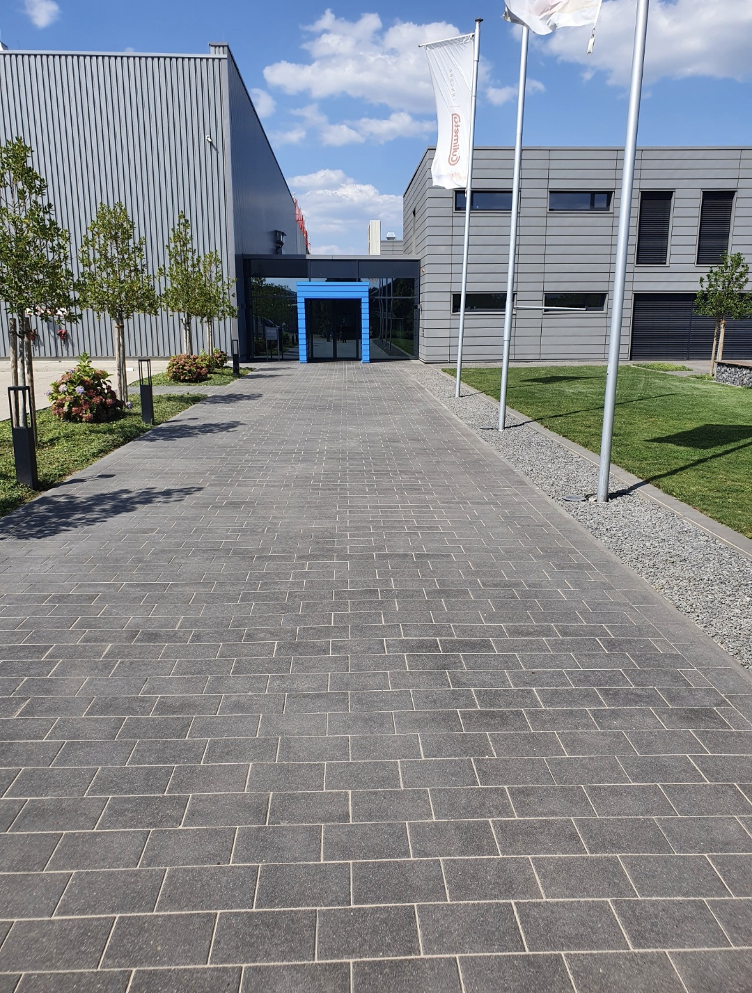 Gepflasterter Weg zu einem modernen grauen Gebäude mit blauen Türen und Fahnen im Wind