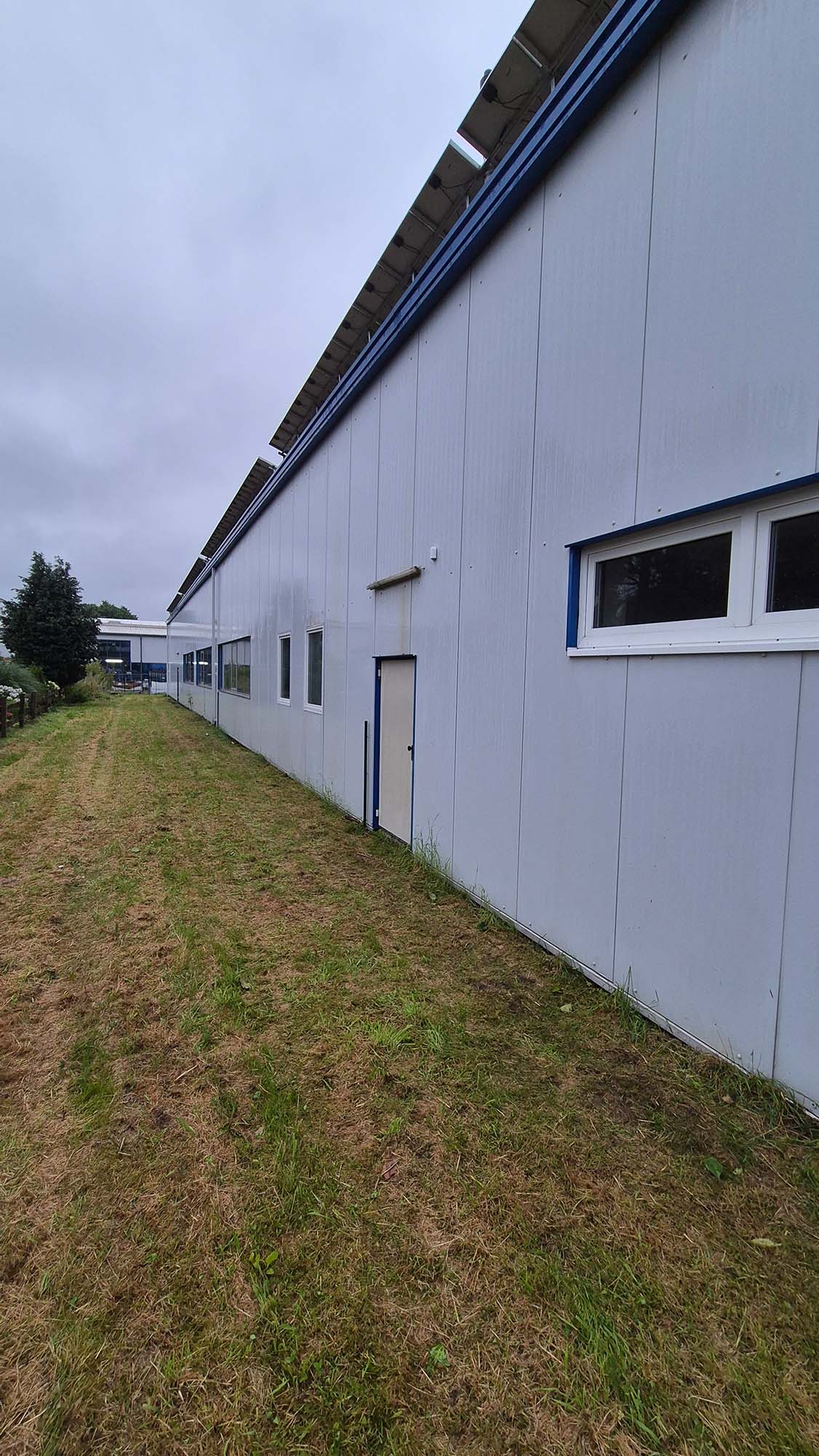 Eine lange graue Industriehalle mit blauen Fensterrahmen und einer Tür im Vordergrund