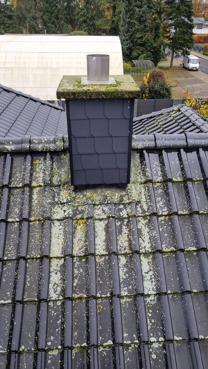 Ein Schornstein auf einem schwarzen Ziegeldach mit Moos und Algenbewuchs