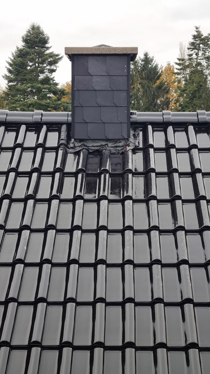 Ein grauer Schornstein steht auf einem glänzenden schwarzen Ziegeldach vor Bäumen