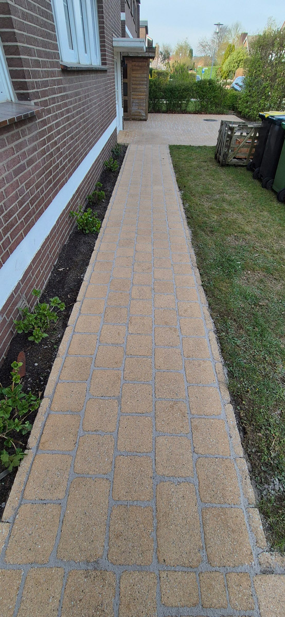 Ein gepflasterter Gehweg mit hellen quadratischen Steinen verläuft entlang eines Hauses