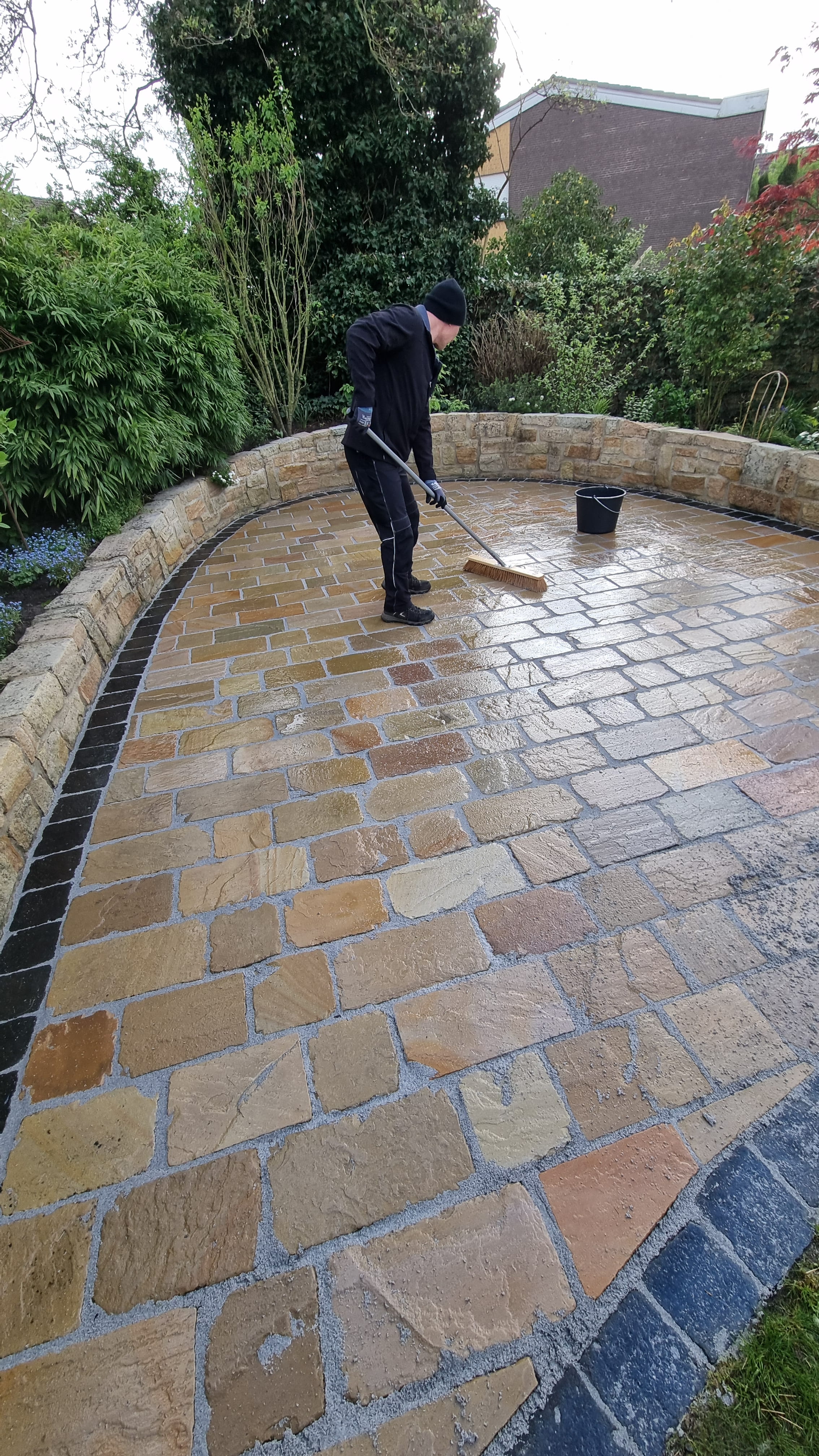 Eine Person mit dunkler Kleidung und Mütze fegt lächelnd einen nassen Steinweg