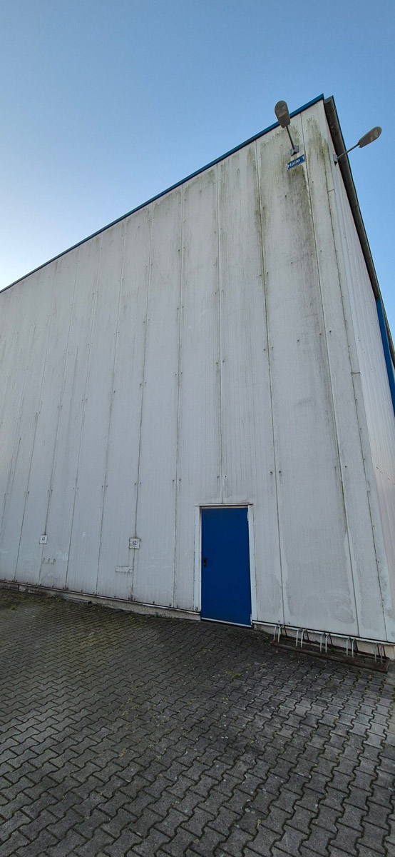 Eine große weiße Hallenwand mit einer kleinen blauen Tür und hohen Lampen oben