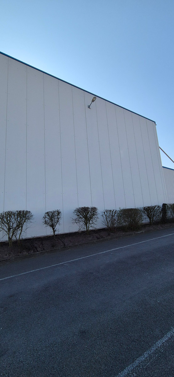 Eine große weiße Wand einer Industriehalle mit Reihen kleiner, kahler Büsche davor